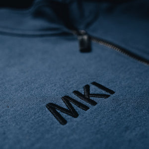 Detail of embroidery on a navy blue fleece bomber jacket with a heavy-duty zipper and distressed leather pull, featuring large pockets with metal grommets, and Shelby Cobra branding patches on the sleeves.
