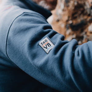 Sleeve patch detail on a navy blue fleece bomber jacket with a heavy-duty zipper and distressed leather pull, featuring large pockets with metal grommets, and Shelby Cobra branding patches on the sleeves.